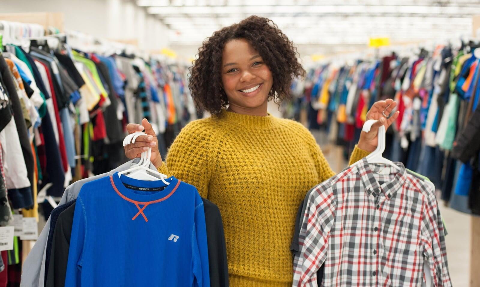 Smiling woman in a gold sweater is shopping and holding up clothing on hangers in both hands.