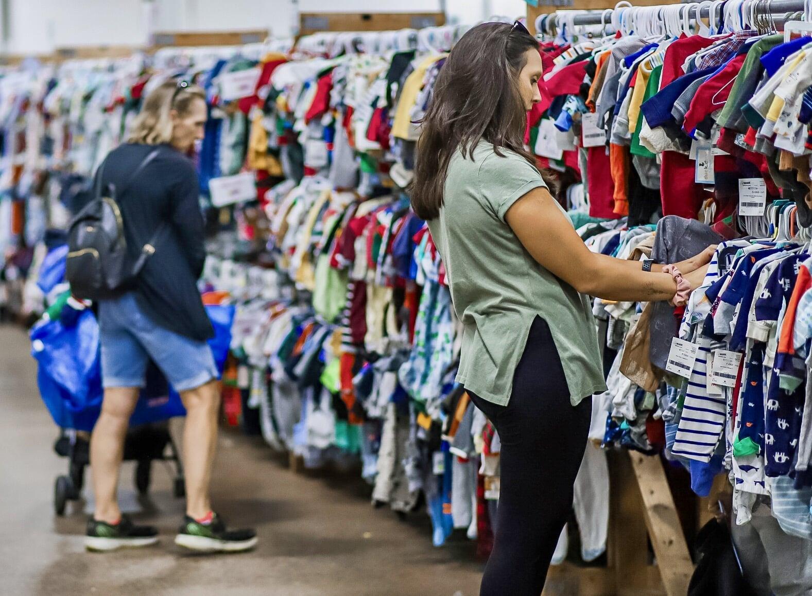 Woman in a grey long sleeve shirt is shopping with her son in the book area of the JBF sale. Her son has a blue striped shirt on and is wearing white and pink bunny ears.