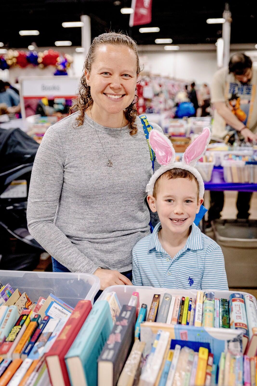 Woman in a grey long sleeve shirt is shopping with her son in the book area of the JBF sale. Her son has a blue striped shirt on and is wearing white and pink bunny ears.