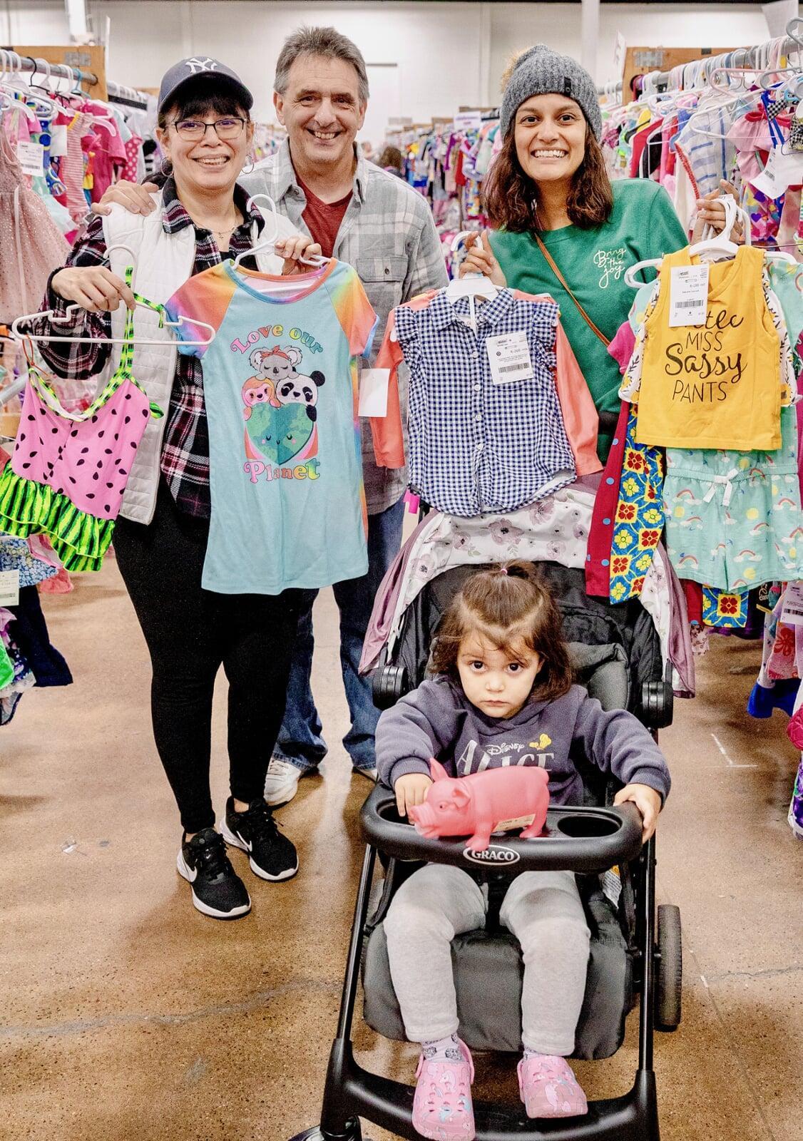 A mom, grandmother and grandfather are shopping with their daughter/granddaughter. The little girl is in a stroller and the adults are standing behind her, all holding up clothing for her.