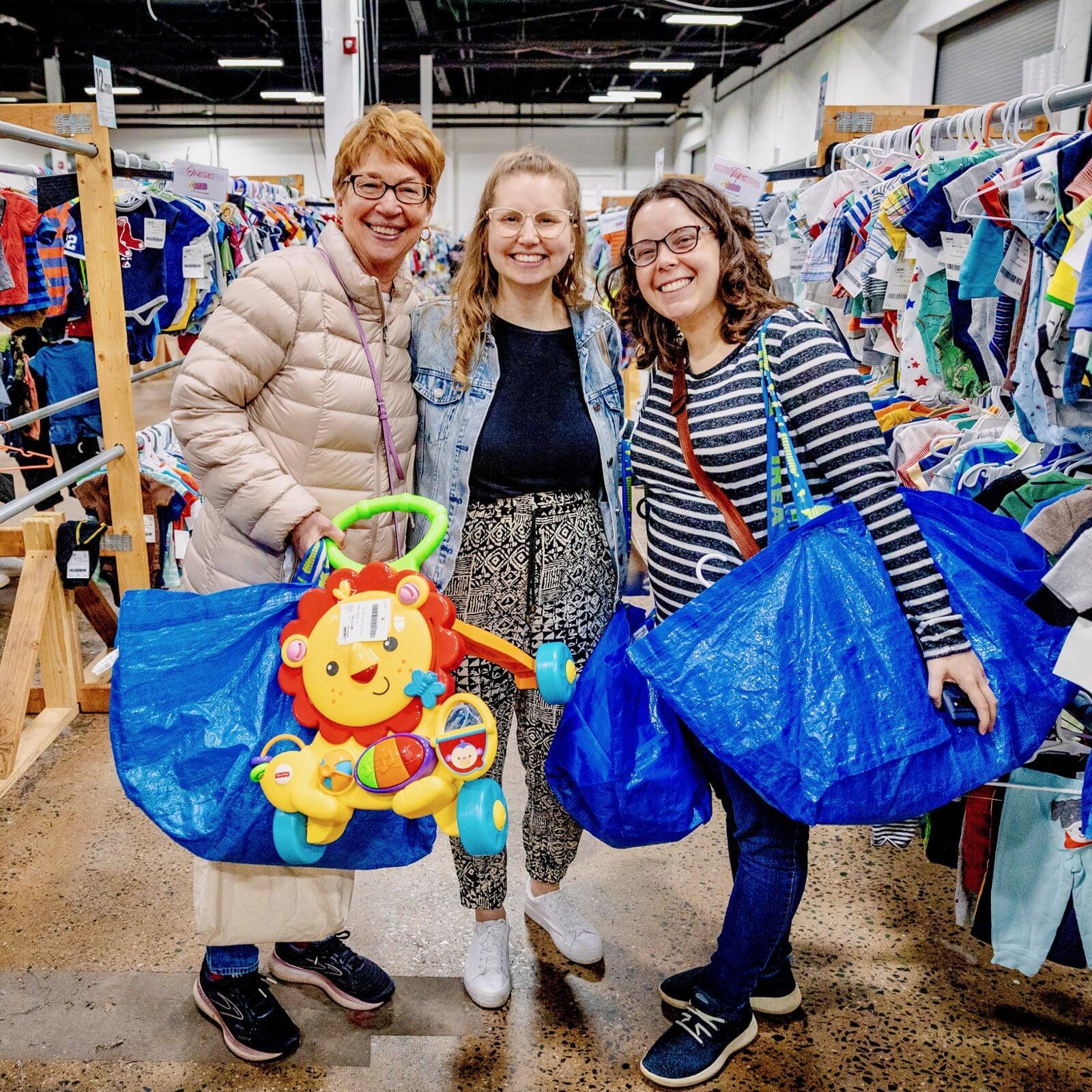 Mother and her two daughters are shopping together. They are standing in between aisles of hanging clothing.