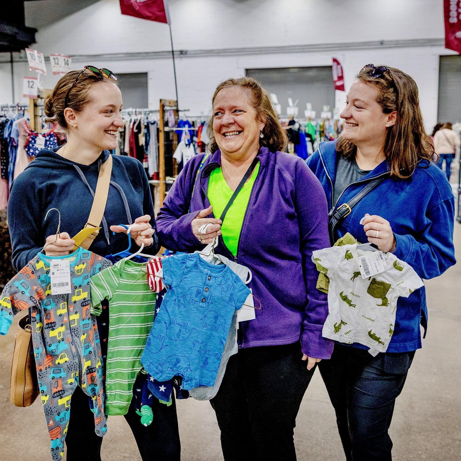 Mother and her two daughters all shopping together. They are all holding baby clothing on hangers.