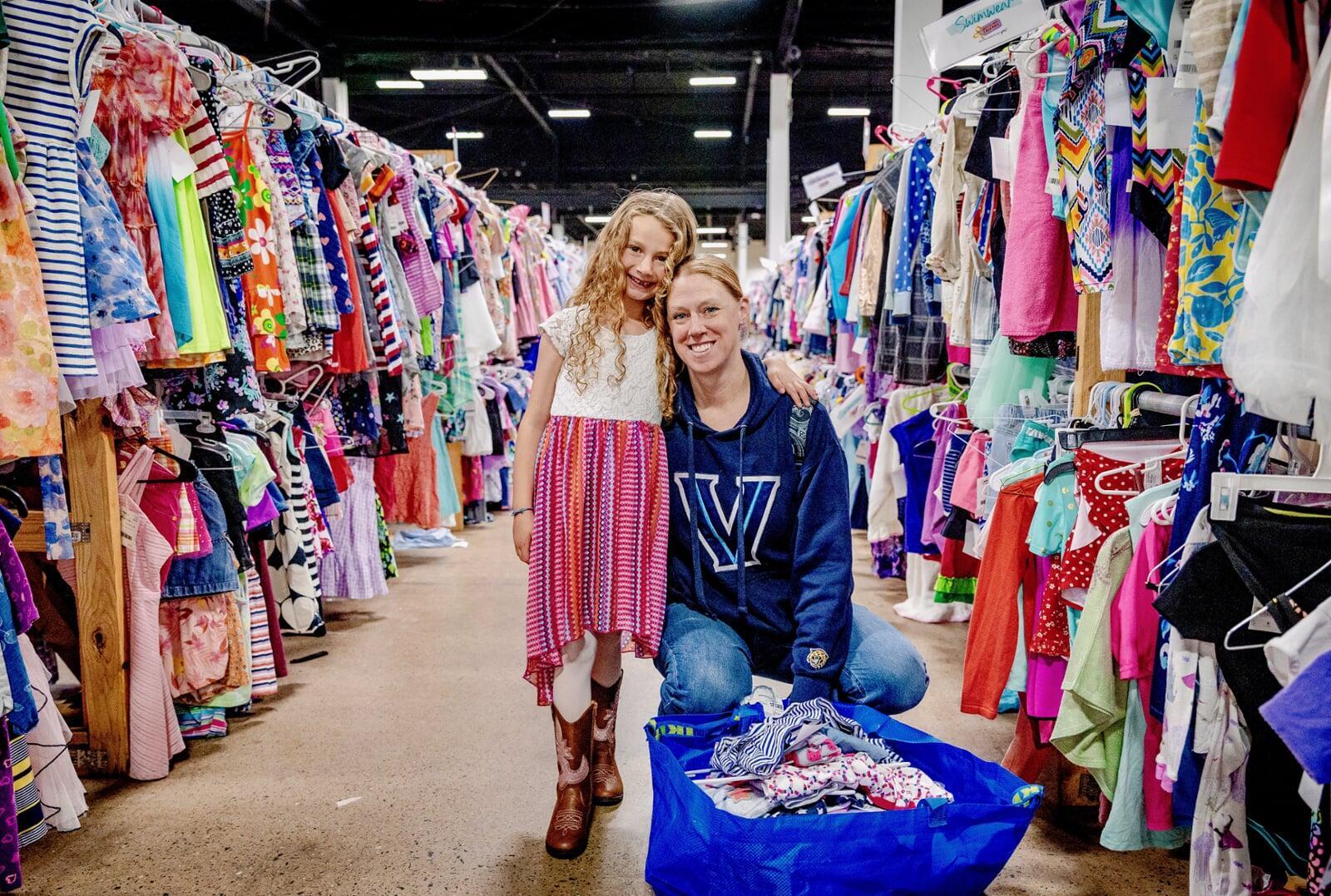 Mom kneels down and has her arm around her daughter. Daughter is standing with her arm around her mom. Mom is wearing a blue Villanova hoodie and daughter is wearing a white, red, purple and orange dress with cowgirl boots. They are in the middle of two aisles of girls clothing and have a bag full of girls clothing to purchase at the JBF sale.