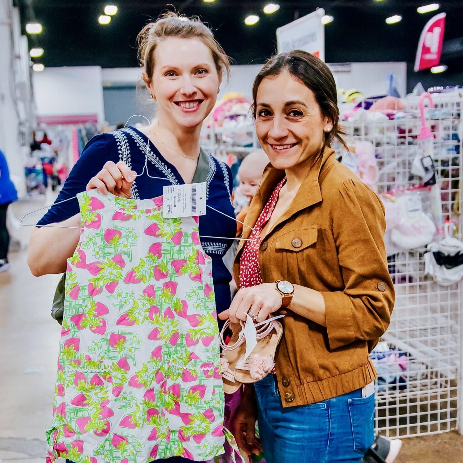 Two women shopping together. One is holding up a sleeveless dress with strawberries on it. The other woman is holding a pair of girls sandals.