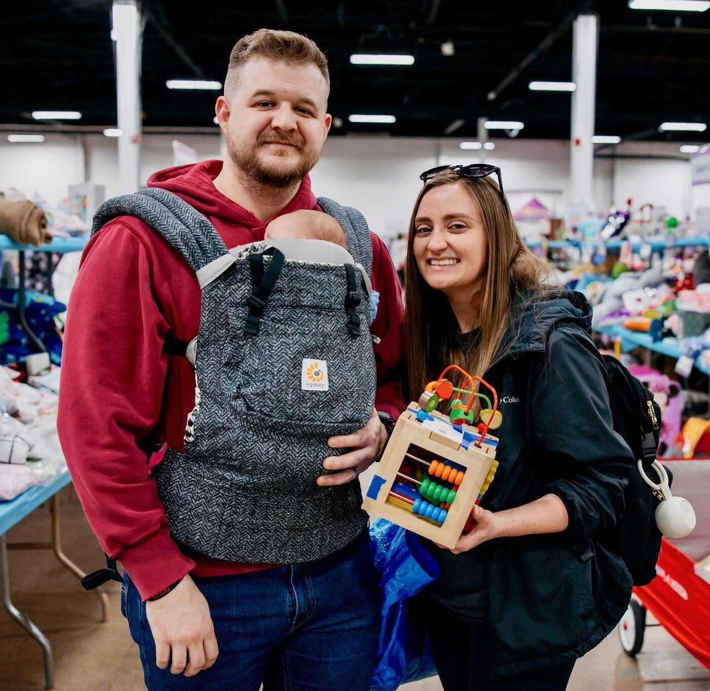 A couple shops at the JBF Oaks sale. Dad is carrying baby in a front carrier and mom is holding an activity cube.