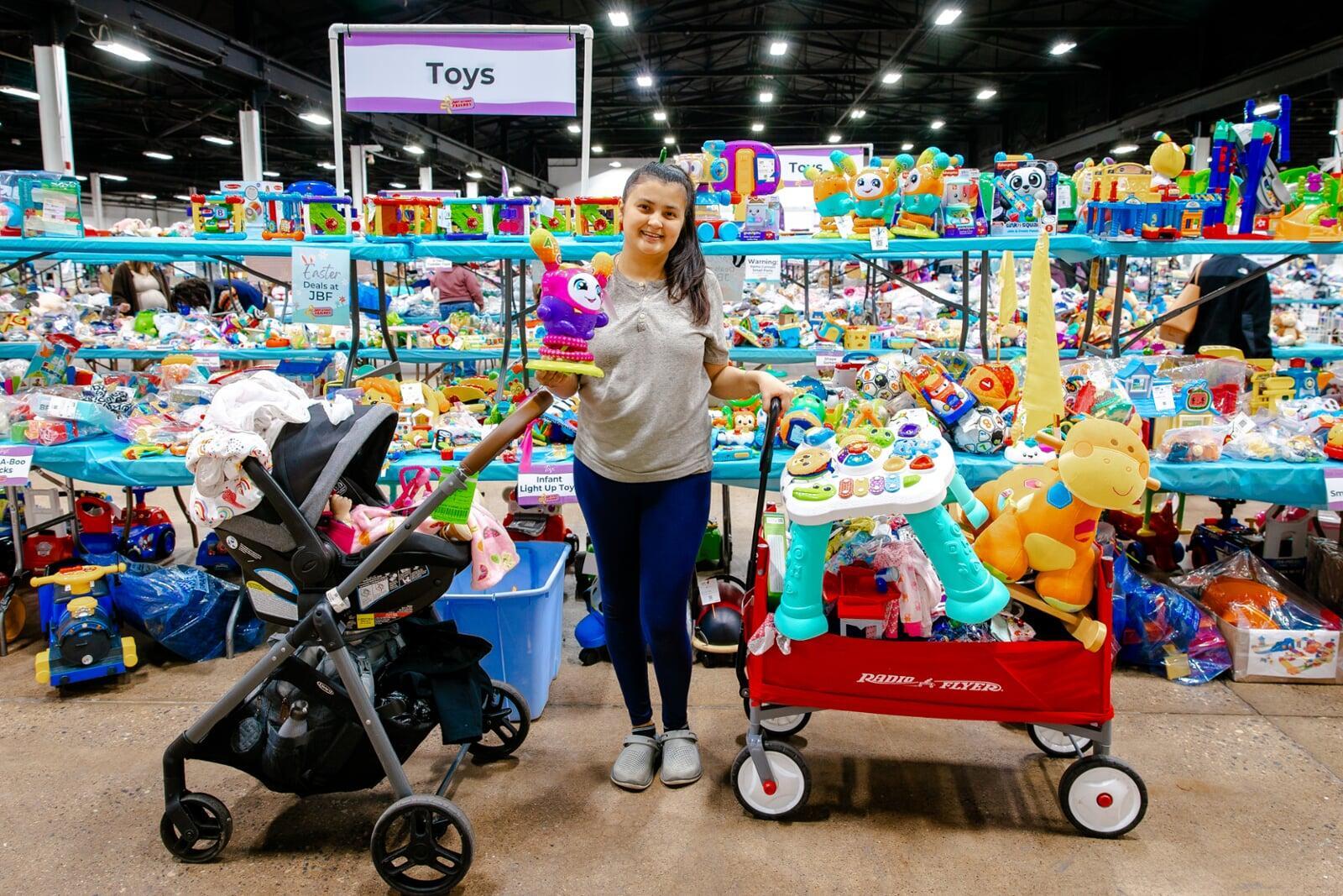 Woman shopping with a baby in a stroller on one side of her and a wagon full of toys on the other side. She is holding up a pink and purple toy.