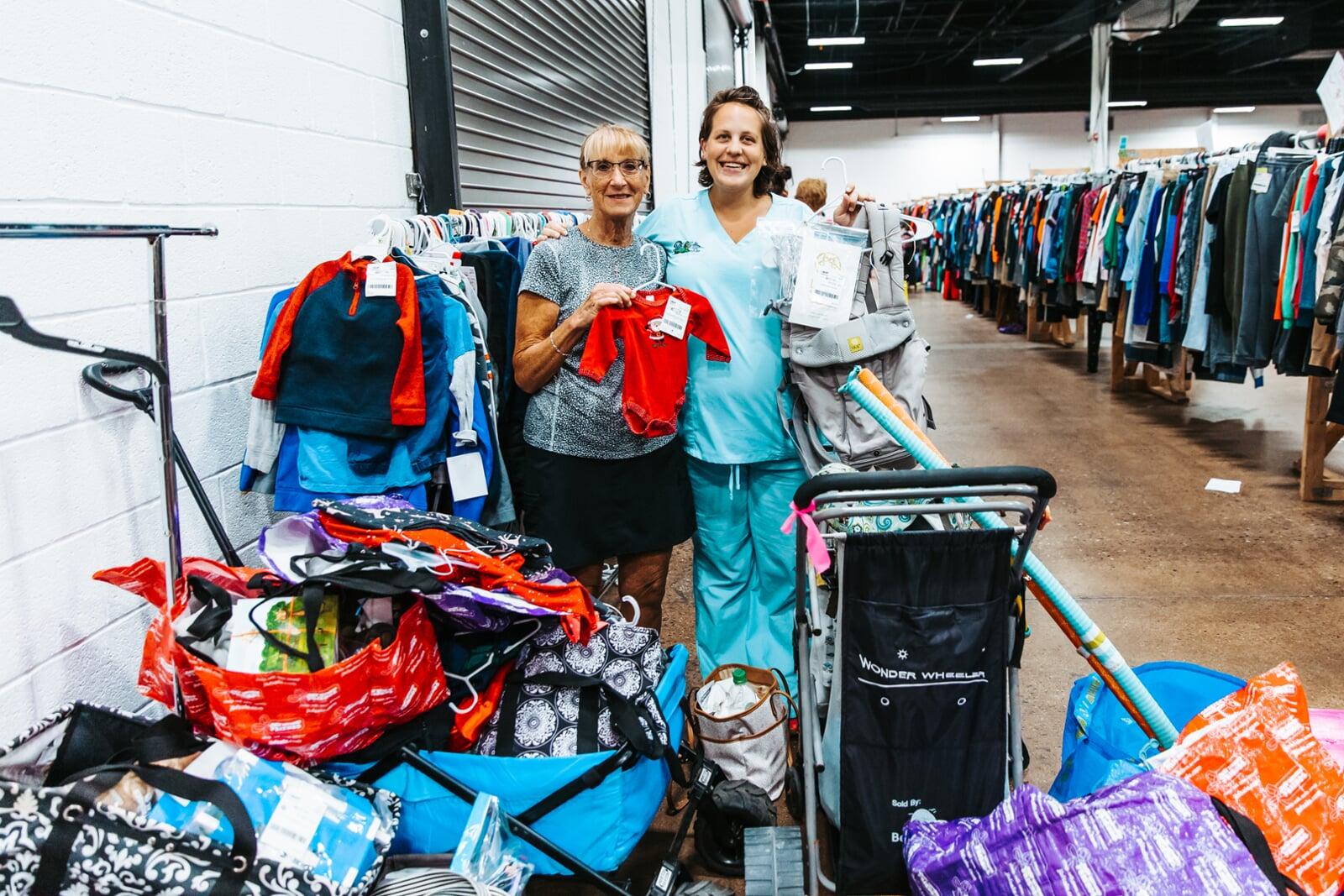 Two women are surrounded by bags, racks and carts of items they found while shopping at the JBF sale.